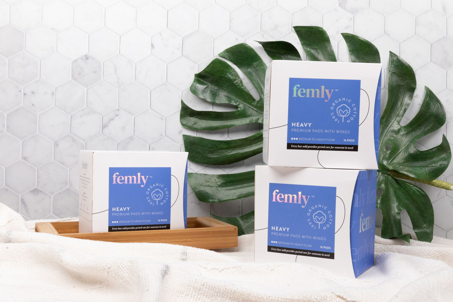 Three boxes of 100% organic cotton heavy menstrual pads in a bathroom setting with a leaf.